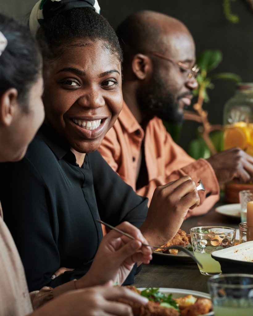 African happy woman talking to girl while sitting at dining table with members of her family